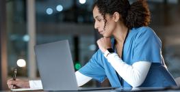 A nurse in blue scrubs sits at a desk, looking at a laptop while writing notes on a clipboard