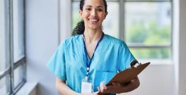 Woman nurse smiling at the camera while holding a clipboard in a hospital hall setting 
