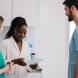 Three healthcare professionals stand together in a clinic. A woman in a white coat is holding a clipboard and smiling while speaking with two colleagues wearing scrubs.